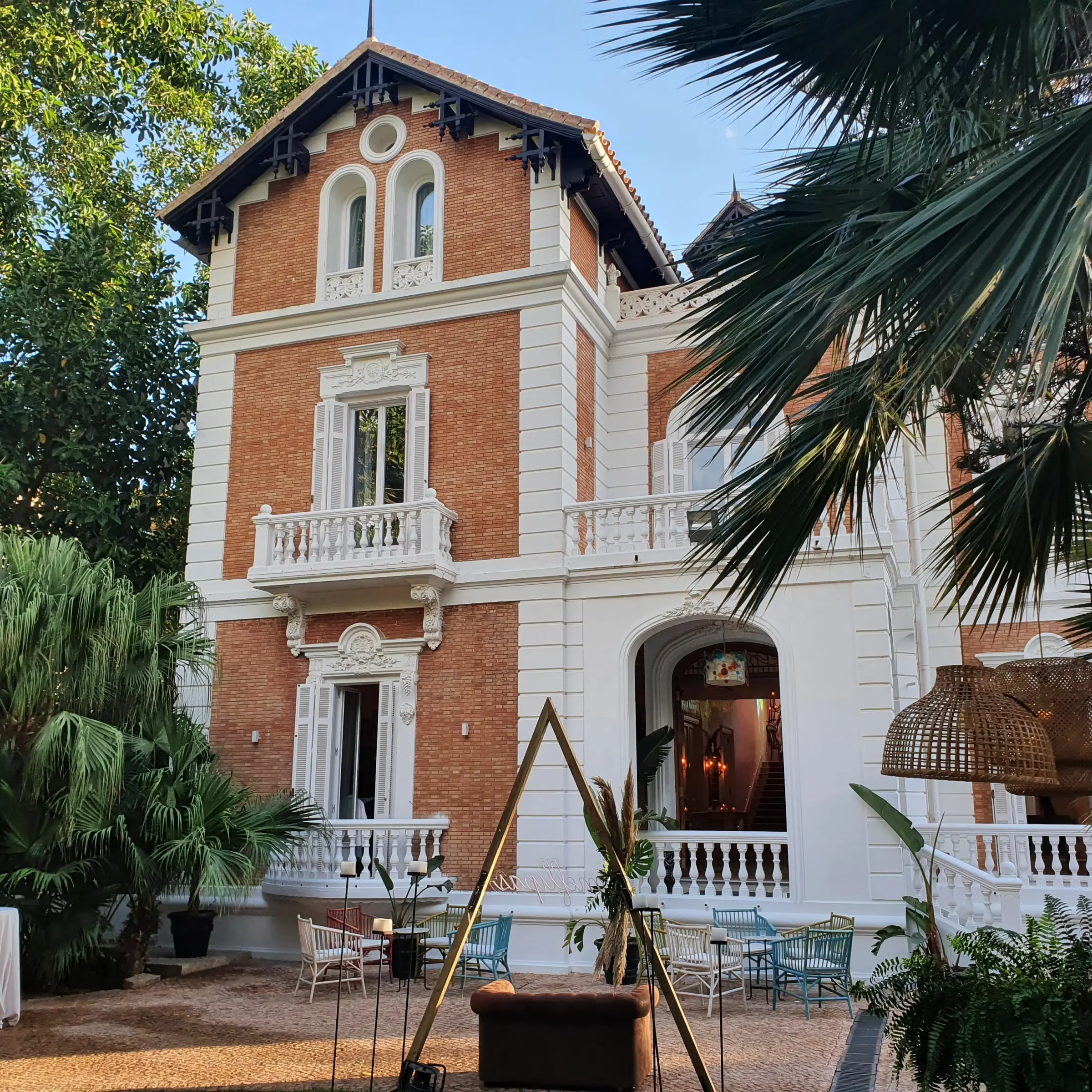 El cuerpo alto del palacio a media tarde: ladrillo, balcones de balaustres blancos y un arco de entrada, con una palmera en primer plano
