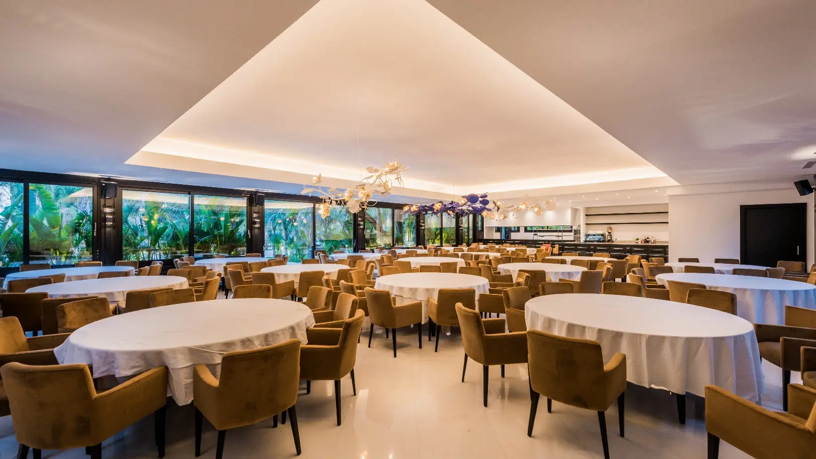 Un comedor acristalado con mesas redondas vestidas de blanco y el jardín tras los ventanales